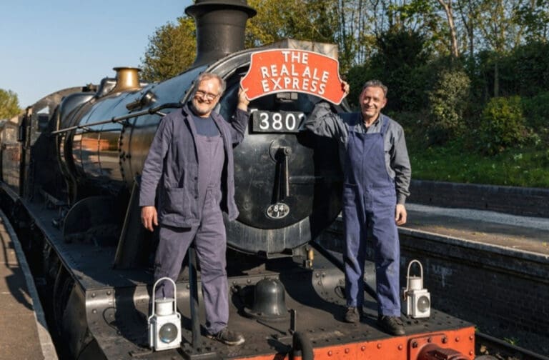 Two railway crew in front of steam locomotive 3802 with a Real Ale Express headboard